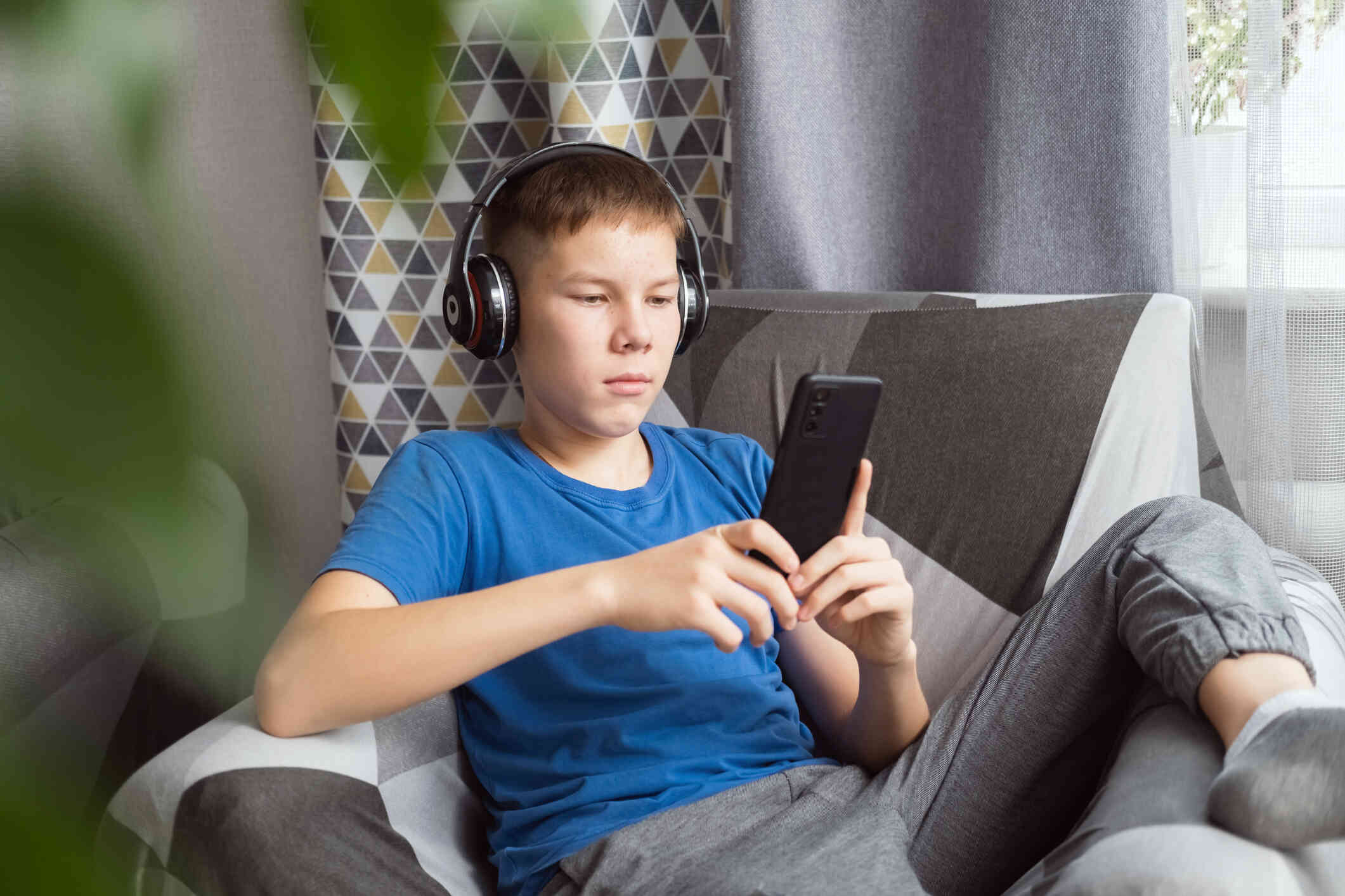 A young boy in a blue shirt sits casually on the couch while wearing over the ear headphones and looking at the cellphone in his hands.